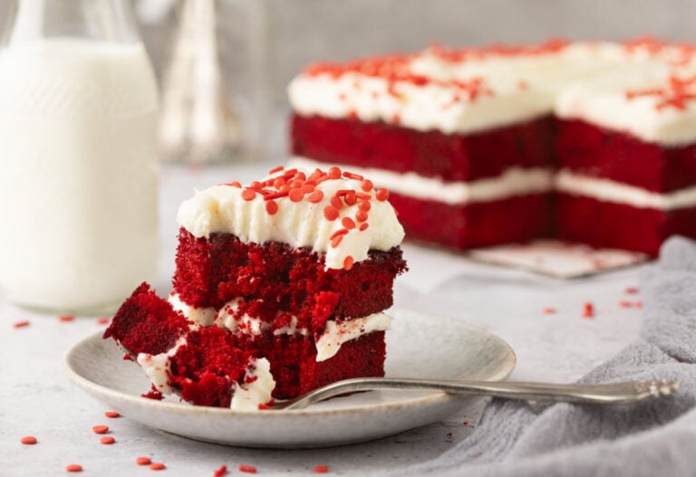 A slice of red velvet cake with white frosting and red sprinkles on a plate with a fork. A bottle of milk and the rest of the cake are in the background.