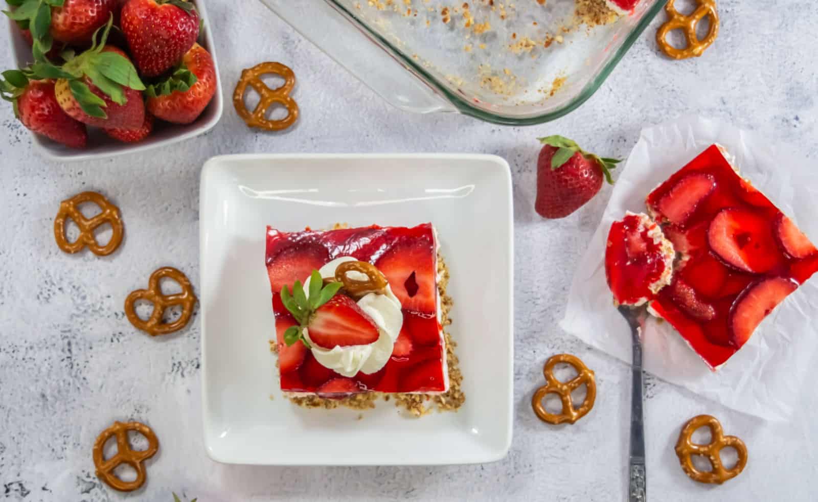 A square slice of strawberry pretzel dessert topped with whipped cream, a strawberry, and a pretzel sits on a white plate, surrounded by more strawberries and pretzels.