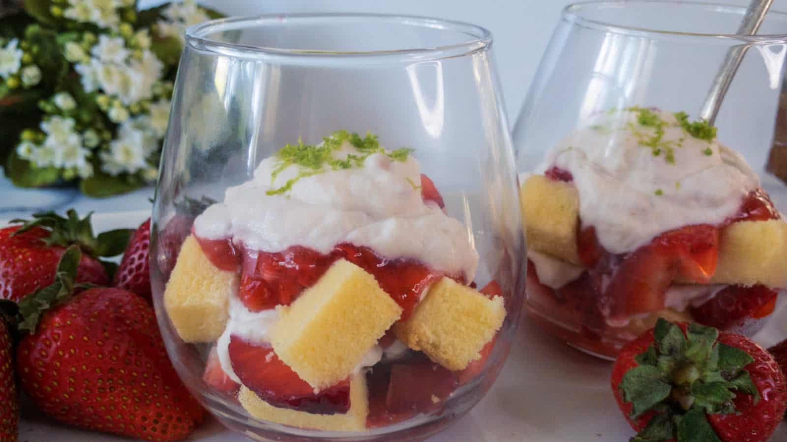 Two shortcake parfaits in glass cups, layered with sponge cake, strawberries, and whipped cream, topped with lime zest. Fresh strawberries and flowers in the background.