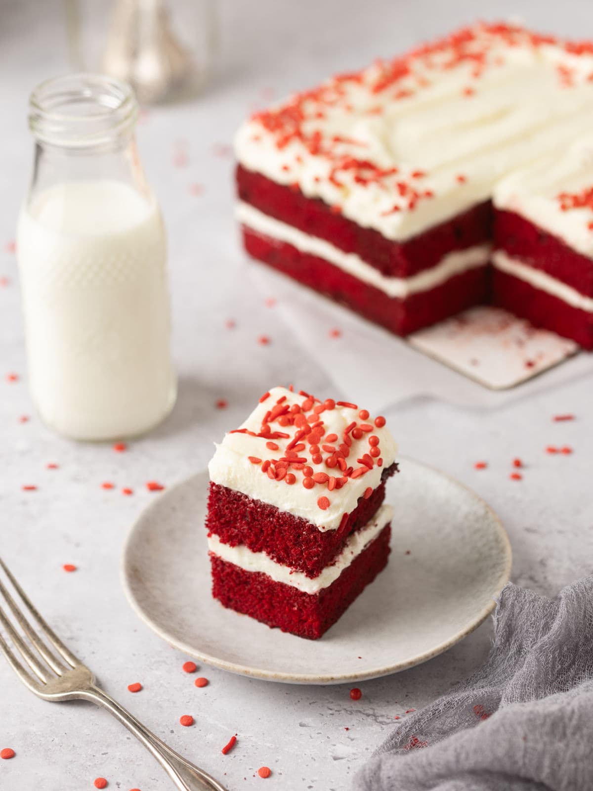 A slice of red velvet cake with cream cheese frosting and red sprinkles on a plate, accompanied by a bottle of milk, a fork, and a larger cake in the background.