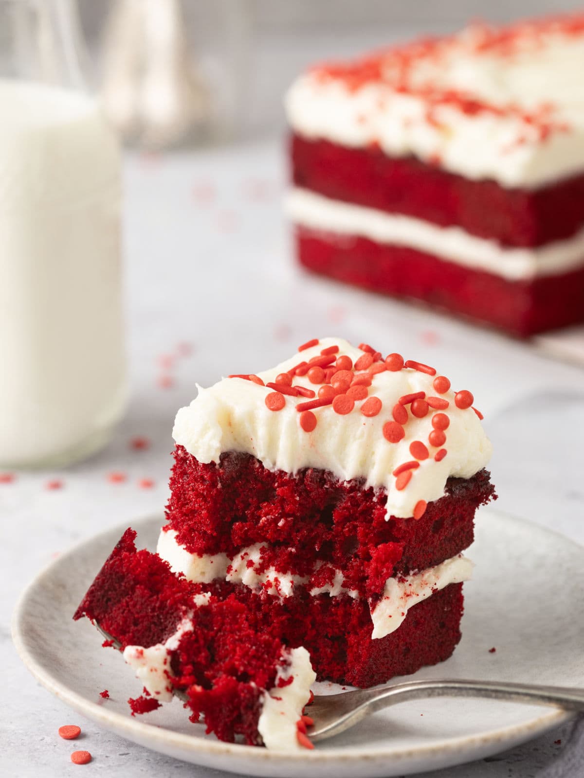 Slice of red velvet cake with cream cheese frosting and red sprinkles on a plate, accompanied by a fork. A bottle of milk and more cake are blurred in the background.