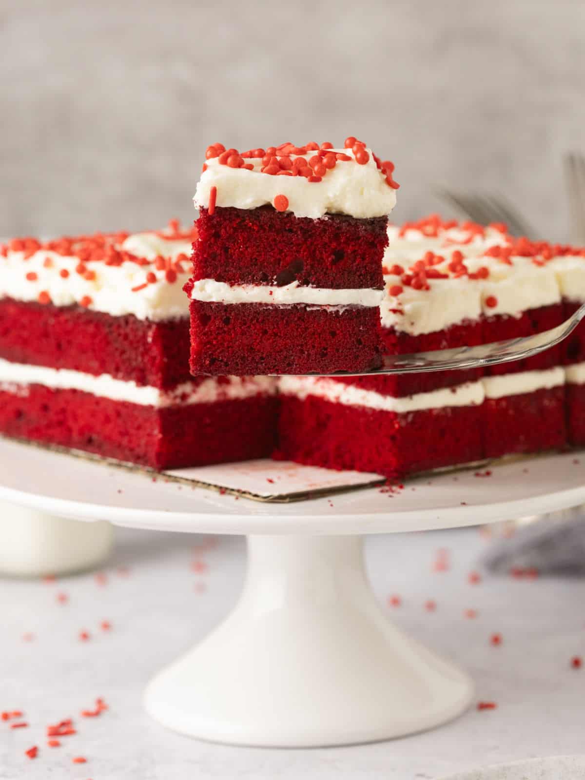 A square slice of red velvet cake with white frosting and red sprinkles is lifted from a cake on a white cake stand.