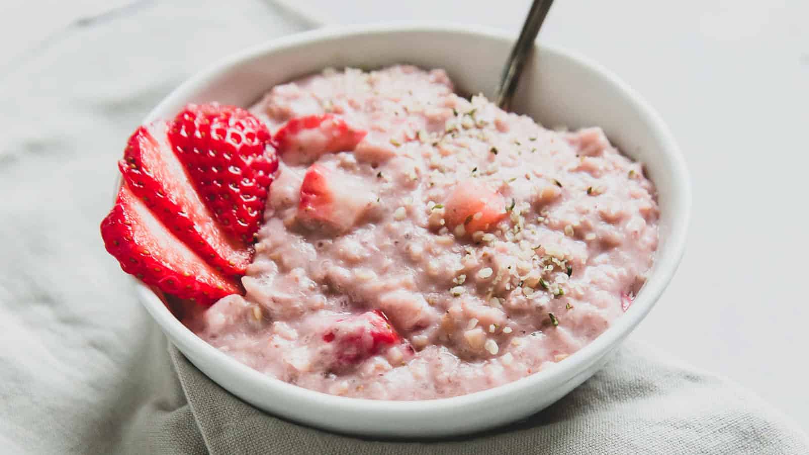 A bowl of strawberry oatmeal topped with sliced strawberries and sprinkled seeds, with a spoon inside the bowl.