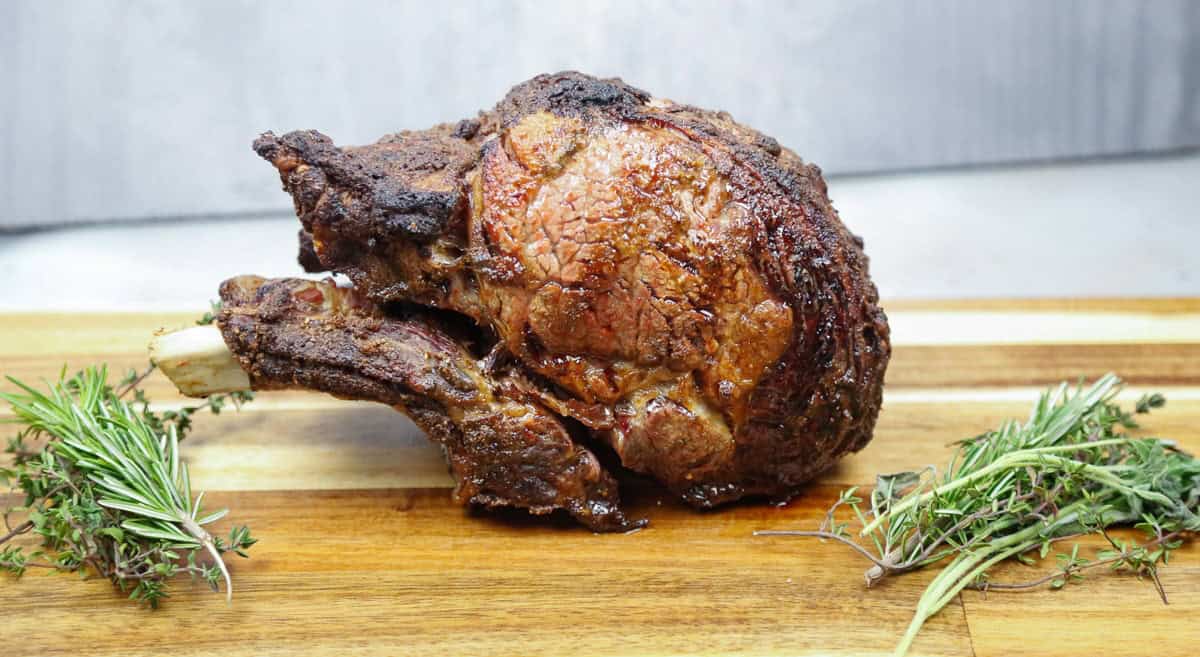 Cooked Tomahawk steak on a wooden board, garnished with fresh rosemary and thyme sprigs.
