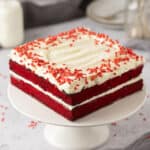 Square red velvet cake with white frosting and red sprinkles on a white cake stand.