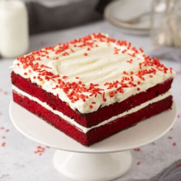 Square red velvet cake with white frosting and red sprinkles on a white cake stand.