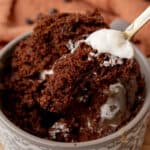 A close-up of a chocolate mug cake with a spoonful topped with whipped cream being lifted from the cup.