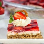 A slice of layered strawberry pretzel dessert topped with whipped cream, a pretzel, and a halved strawberry on a white plate.