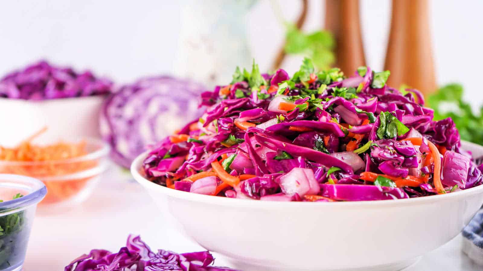 A bowl of vibrant red cabbage salad with carrots and herbs, surrounded by bowls of shredded ingredients on a light background.