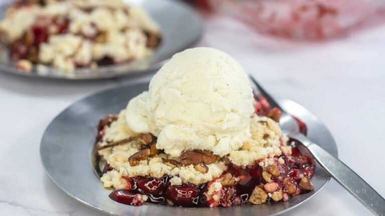 A plate of cherry cobbler topped with a scoop of vanilla ice cream and pecans, with a spoon next to it. Another plate of the same dessert is blurred in the background.