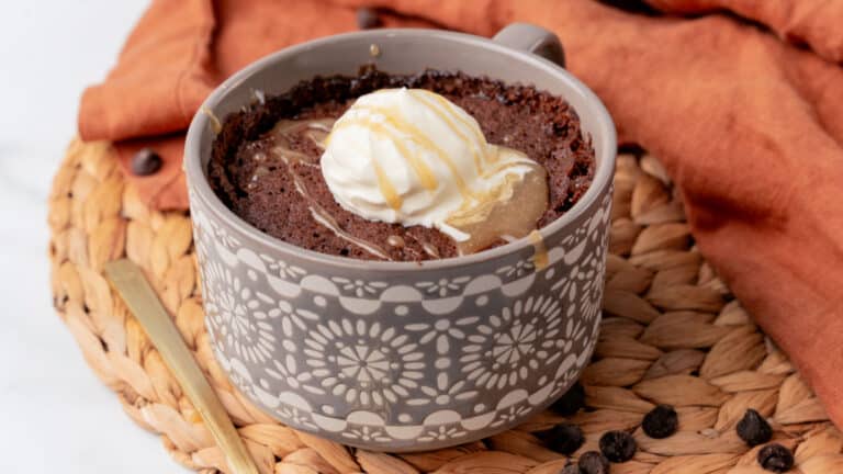 A chocolate mug cake topped with a scoop of vanilla ice cream and caramel drizzle sits on a woven mat next to an orange cloth.