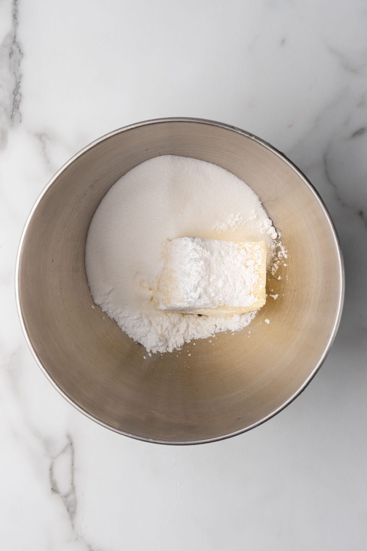 A metal mixing bowl containing granulated sugar, powdered sugar, and a block of butter on a marble countertop.