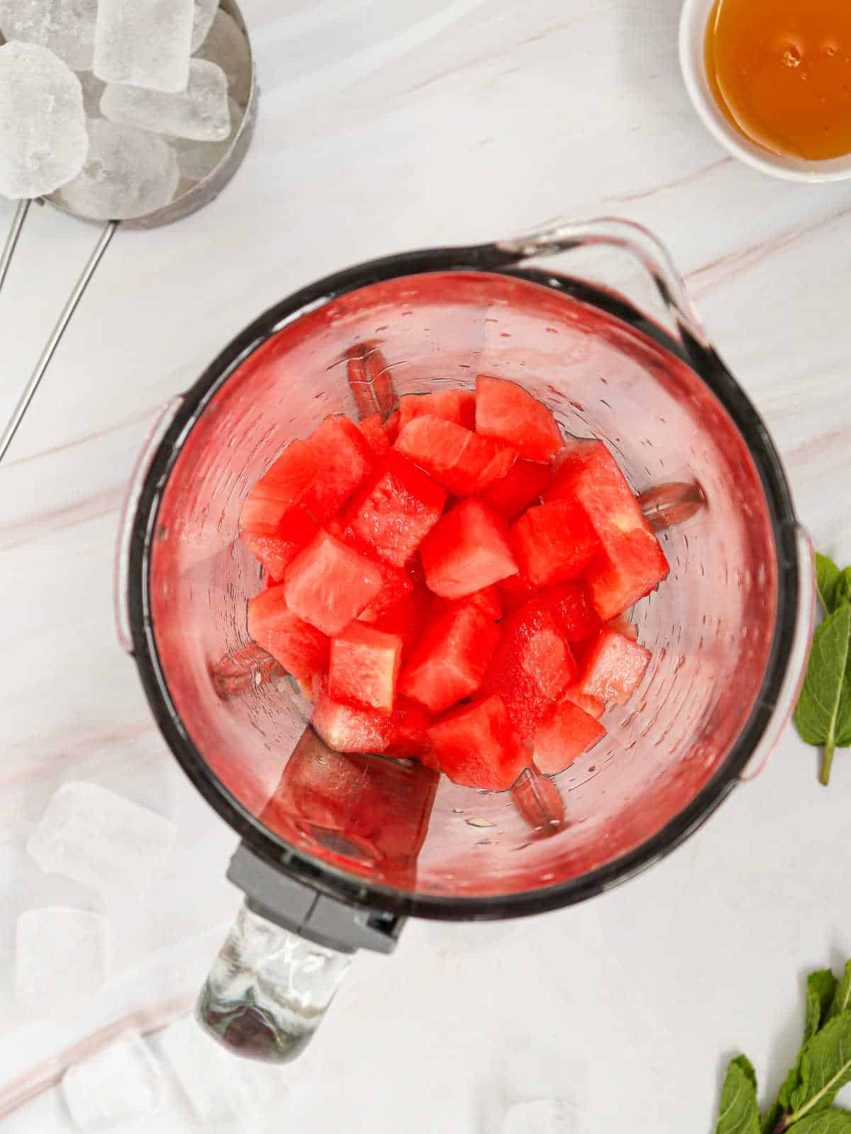 Cubed watermelon pieces inside a blender jug on a white surface, surrounded by ice cubes, a bowl of honey, and mint leaves.
