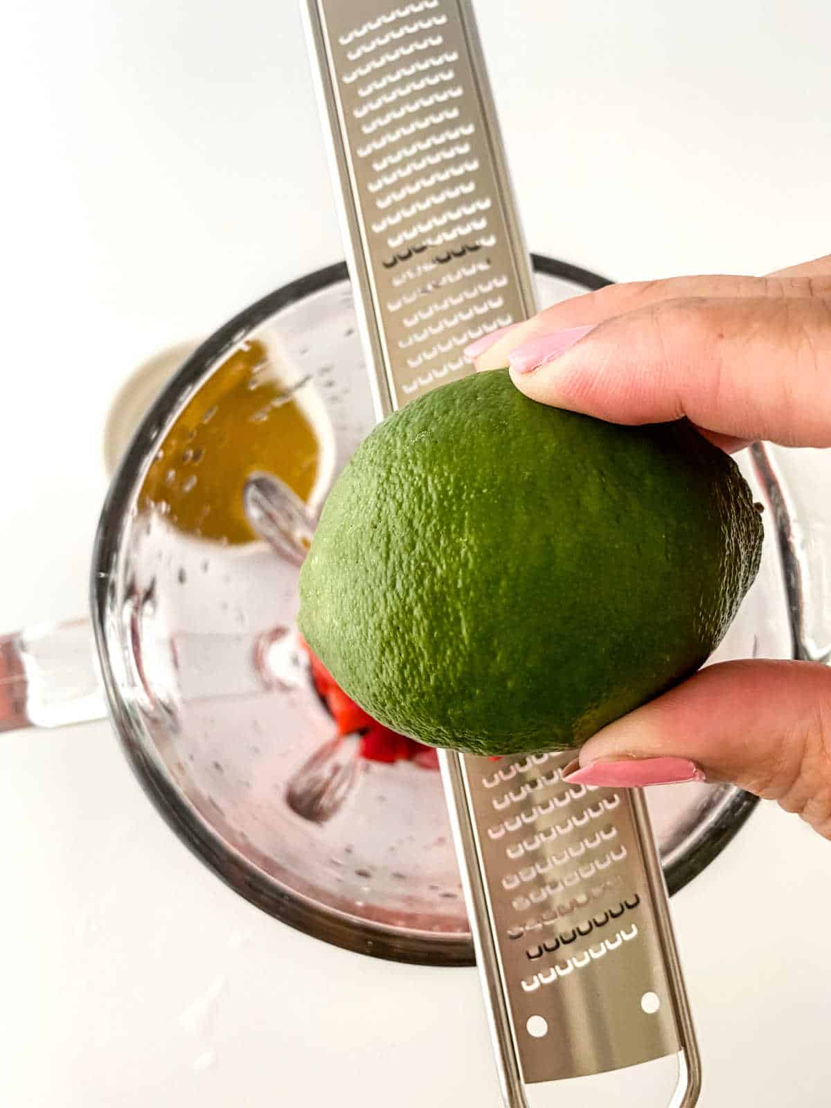 A hand holds a lime over a metal zester placed on a blender, with chopped ingredients visible inside the blender jar.