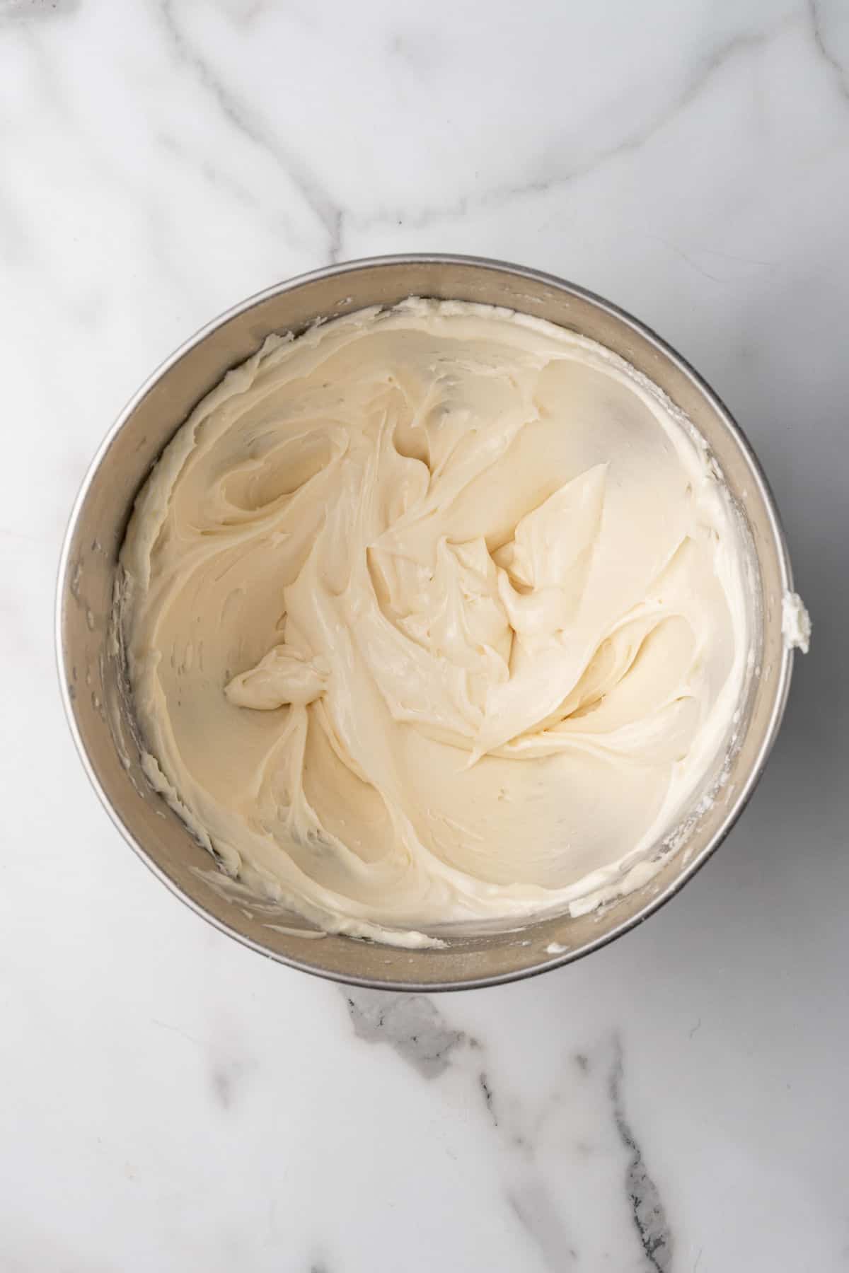 A stainless steel bowl filled with smooth, creamy white frosting sits on a white marble surface.