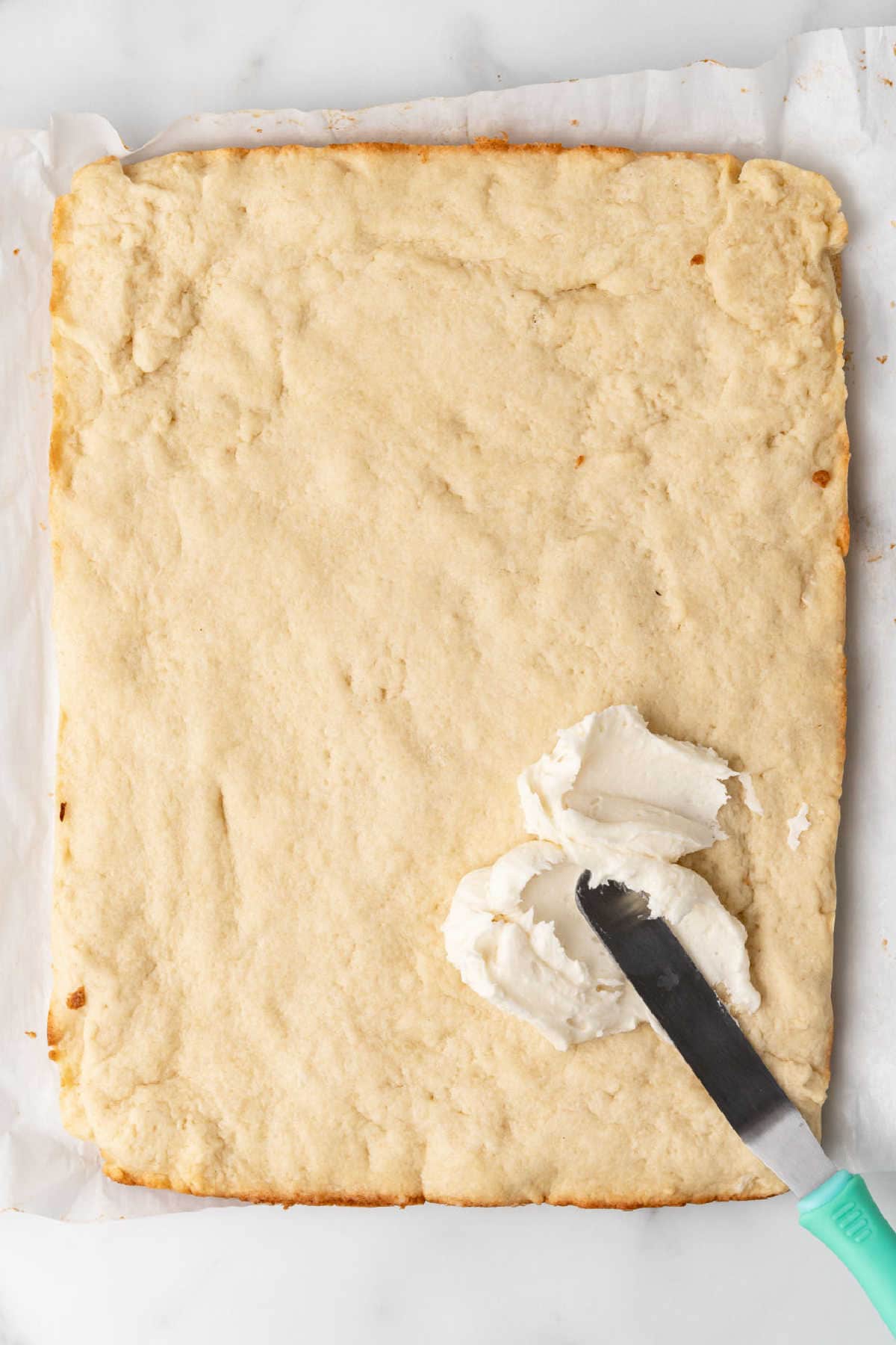 A rectangular sheet of baked cake on parchment paper with a spatula spreading white frosting on one corner.