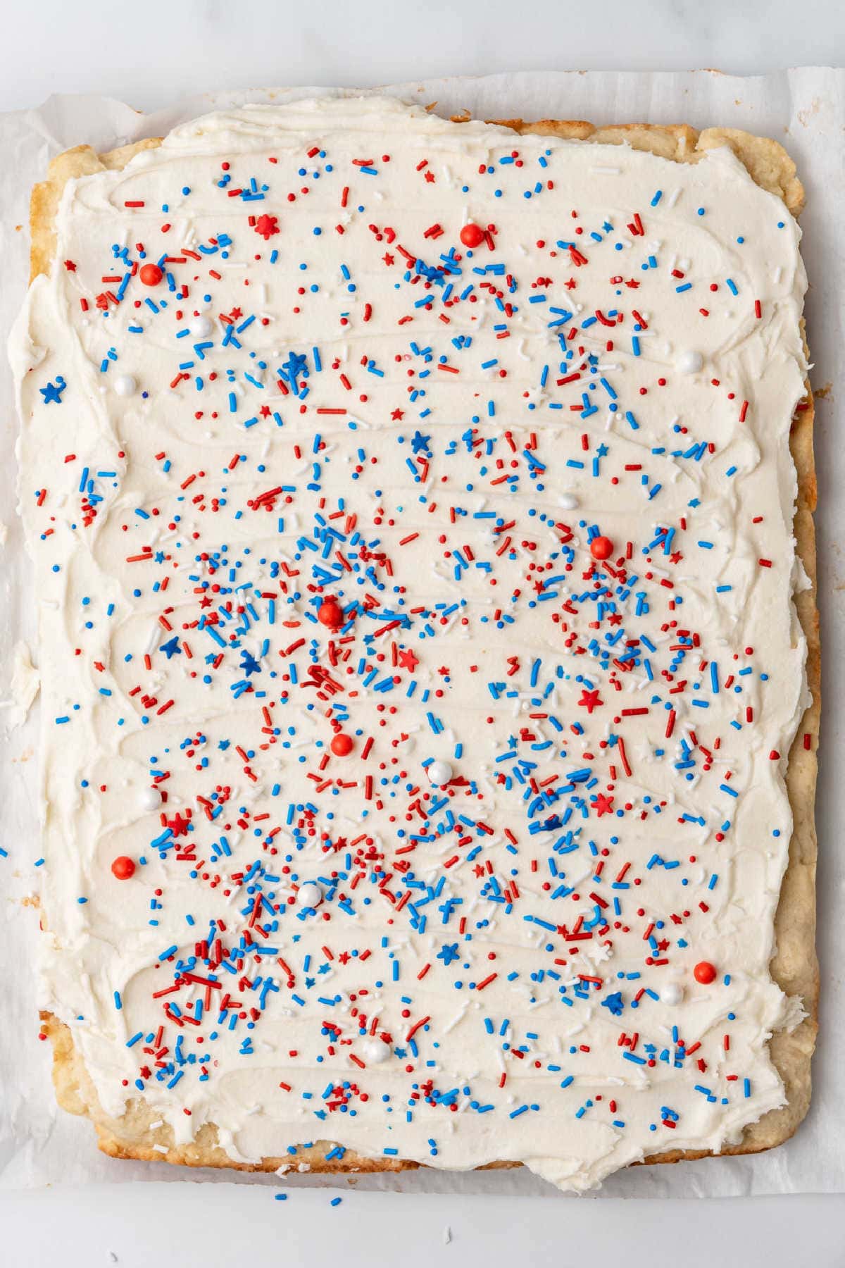 A rectangular cake with white frosting, topped with red, white, and blue sprinkles, on a parchment-lined surface.