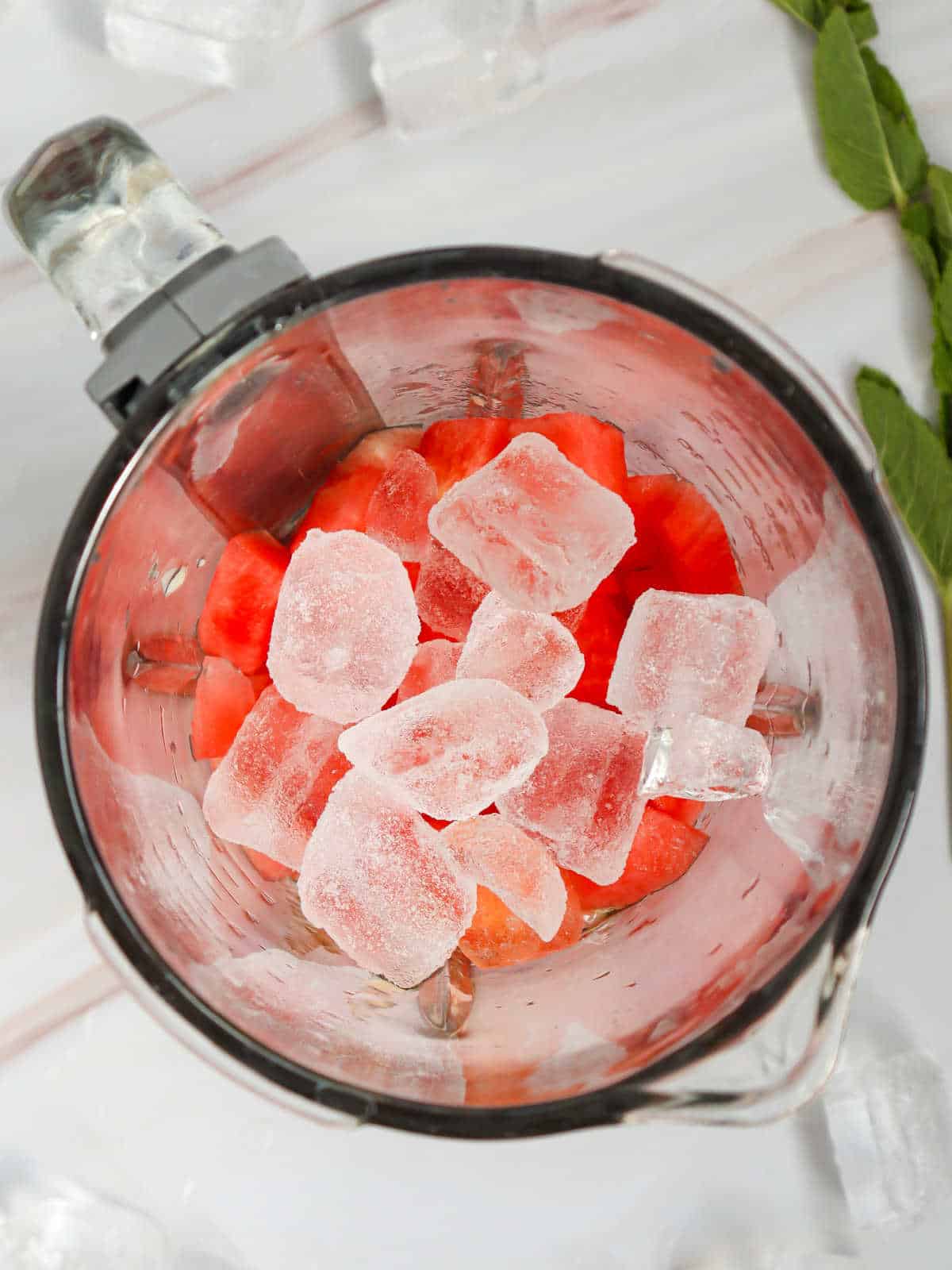 Overhead view of a blender containing watermelon chunks and ice cubes, placed on a light surface with mint leaves and ice nearby.