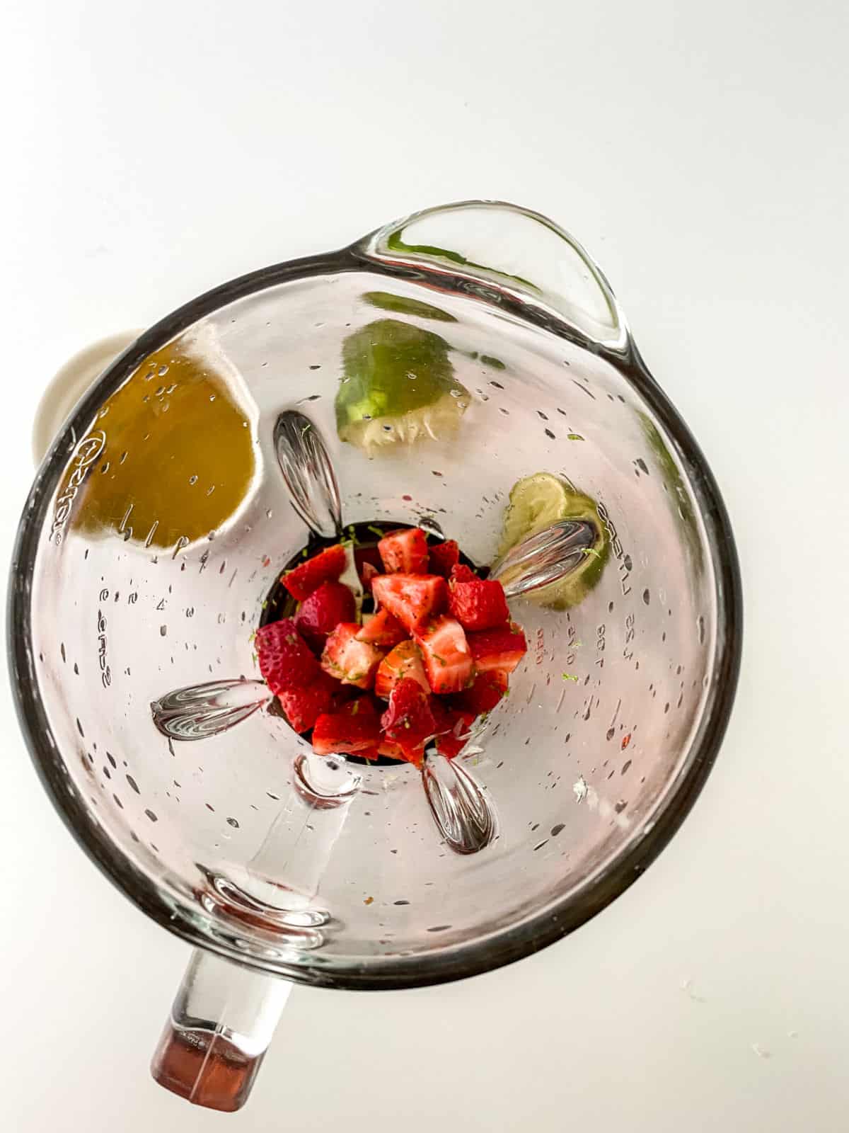 Overhead view of a blender containing chopped strawberries, lime wedges, and an orange slice on a white surface.