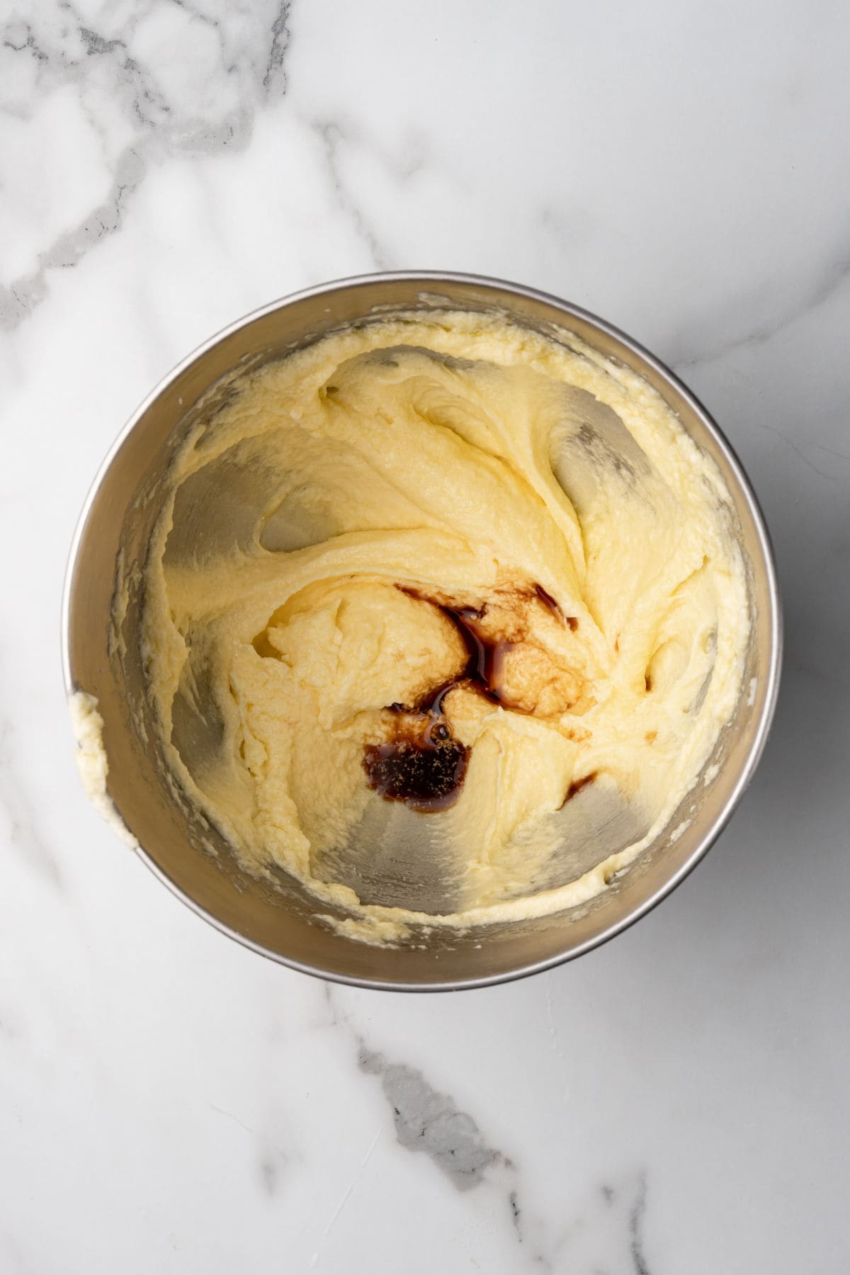 A metal mixing bowl with yellow cake batter and a splash of vanilla extract on top, sitting on a white marble surface.