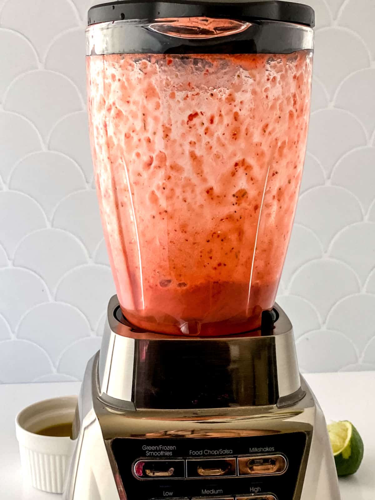 A blender filled with a pink smoothie mixture is blending on a countertop. A small white cup and a lime wedge are nearby.