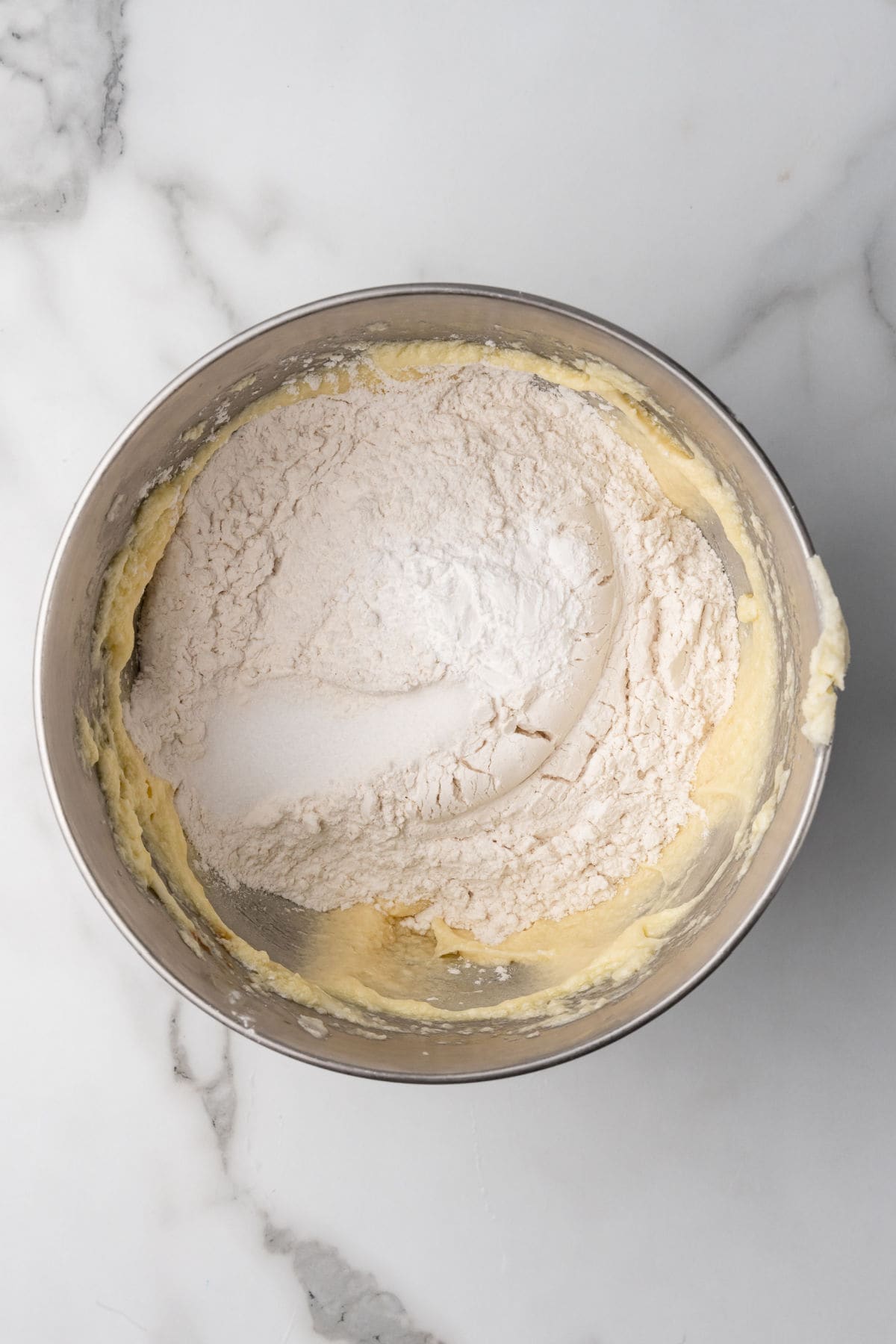 A metal mixing bowl containing creamed butter and sugar with flour and baking powder added on top, ready to be mixed, sits on a marble surface.