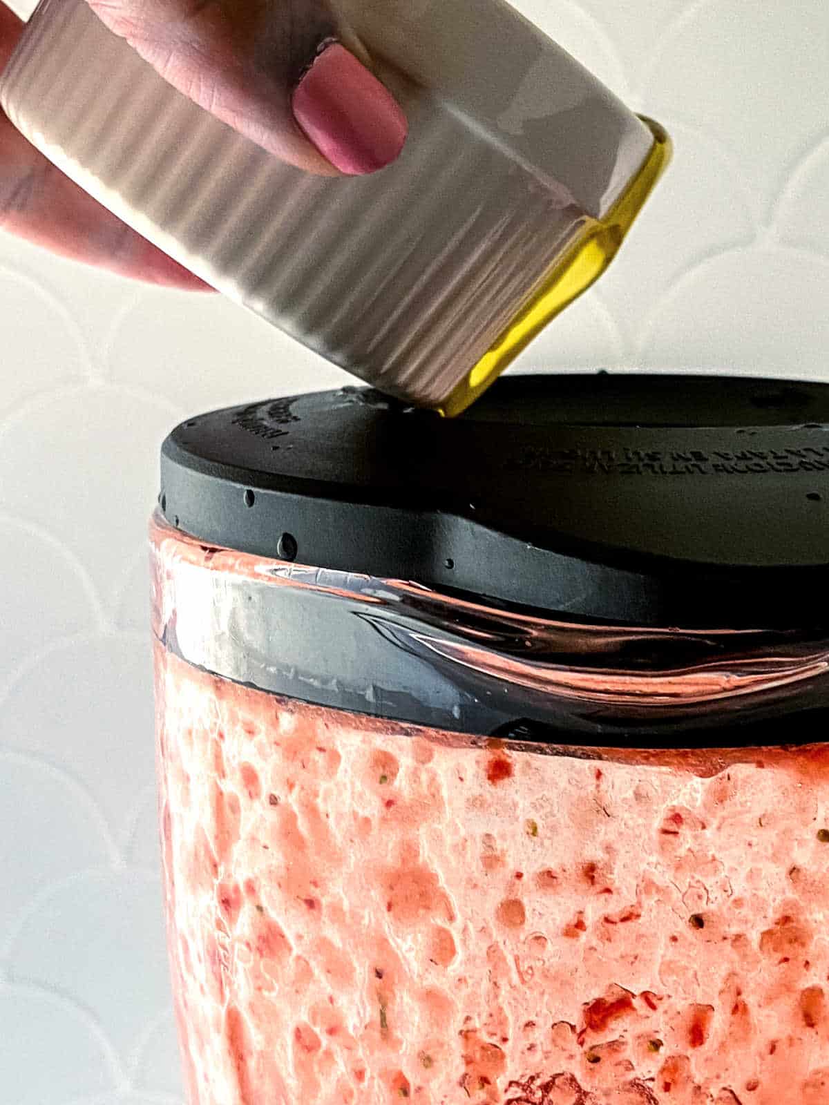 A hand pours olive oil from a small white container onto the black lid of a glass jar filled with bubbly pink dough.