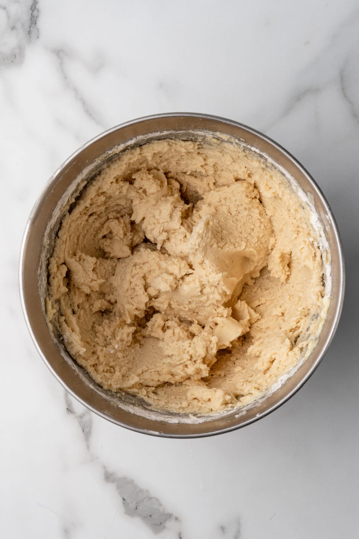 A stainless steel bowl filled with creamy, light-colored cookie dough sits on a white marble surface.