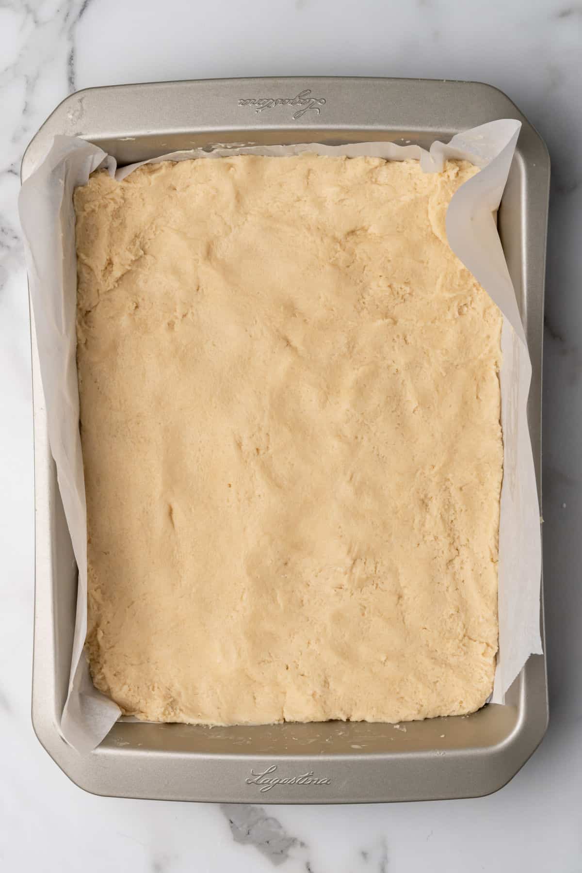 A rectangular baking pan lined with parchment paper and filled with an even layer of unbaked dough, set on a marble surface.