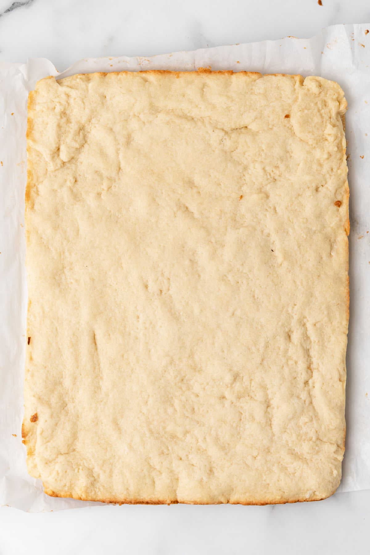A rectangular baked shortbread crust on parchment paper, viewed from above on a marble surface.