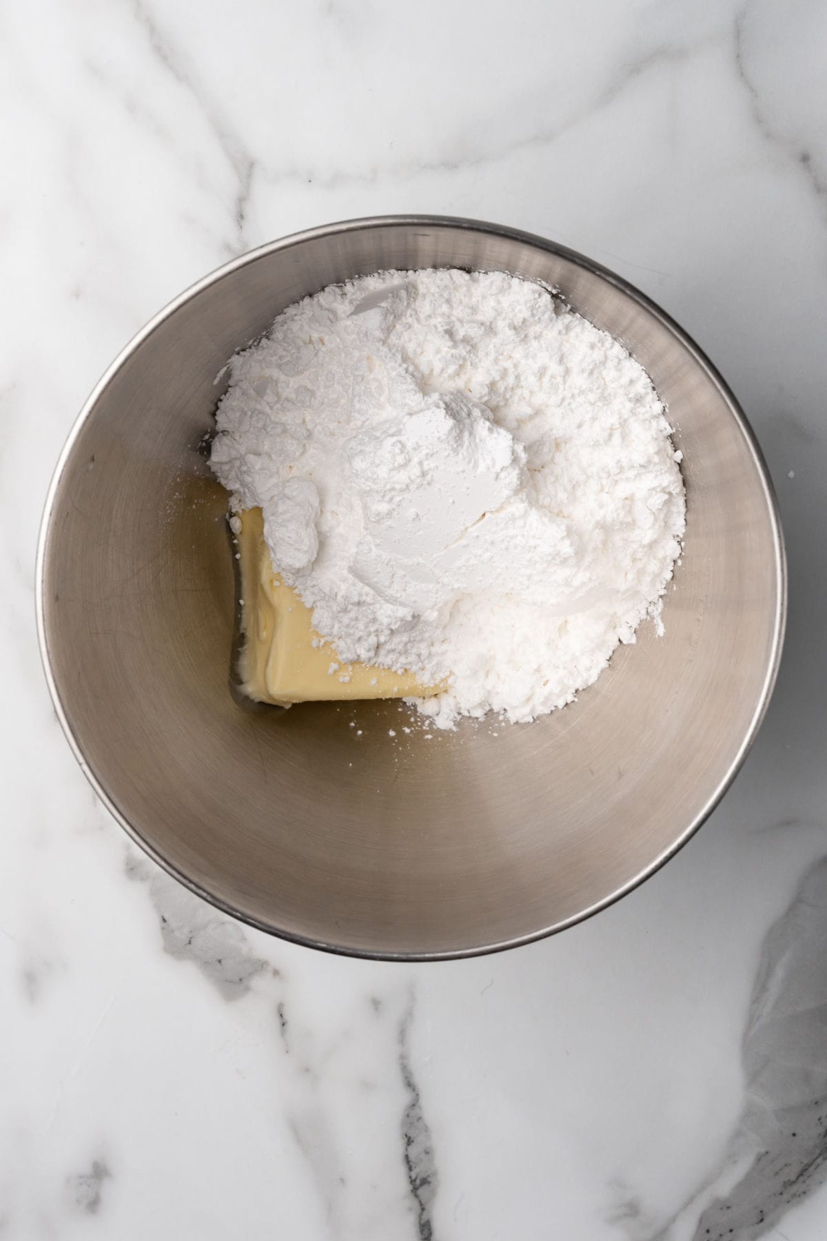 A metal mixing bowl containing a block of butter and a pile of powdered sugar on a white marble countertop.