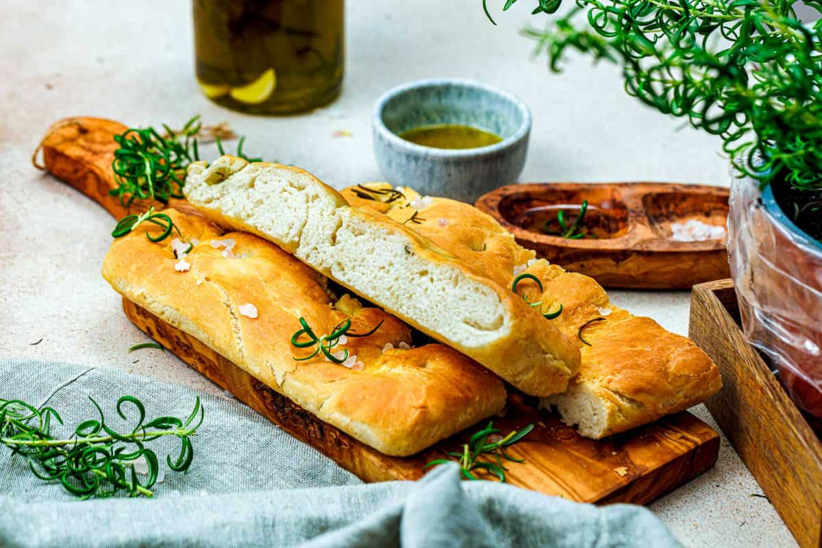 Two pieces of focaccia bread topped with rosemary and sea salt are placed on a wooden board, with a small bowl of olive oil and fresh herbs nearby.