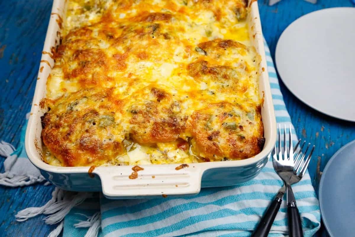 A baked casserole topped with golden melted cheese sits in a rectangular dish on a blue table, next to a striped towel, two forks, and empty plates.