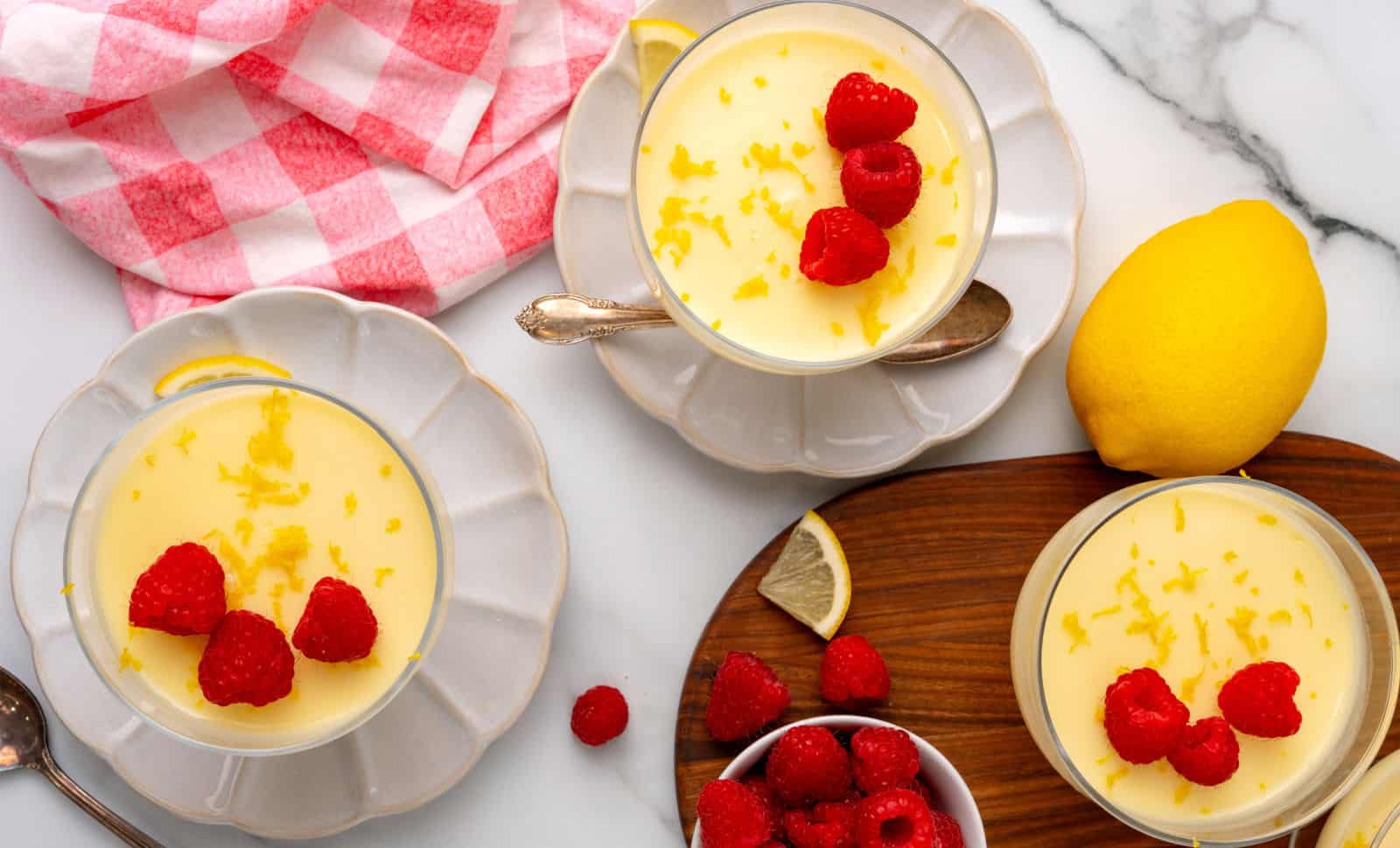 Three bowls of lemon mousse topped with fresh raspberries and lemon zest are arranged on a table with a lemon, loose raspberries, and a red-checkered cloth nearby.