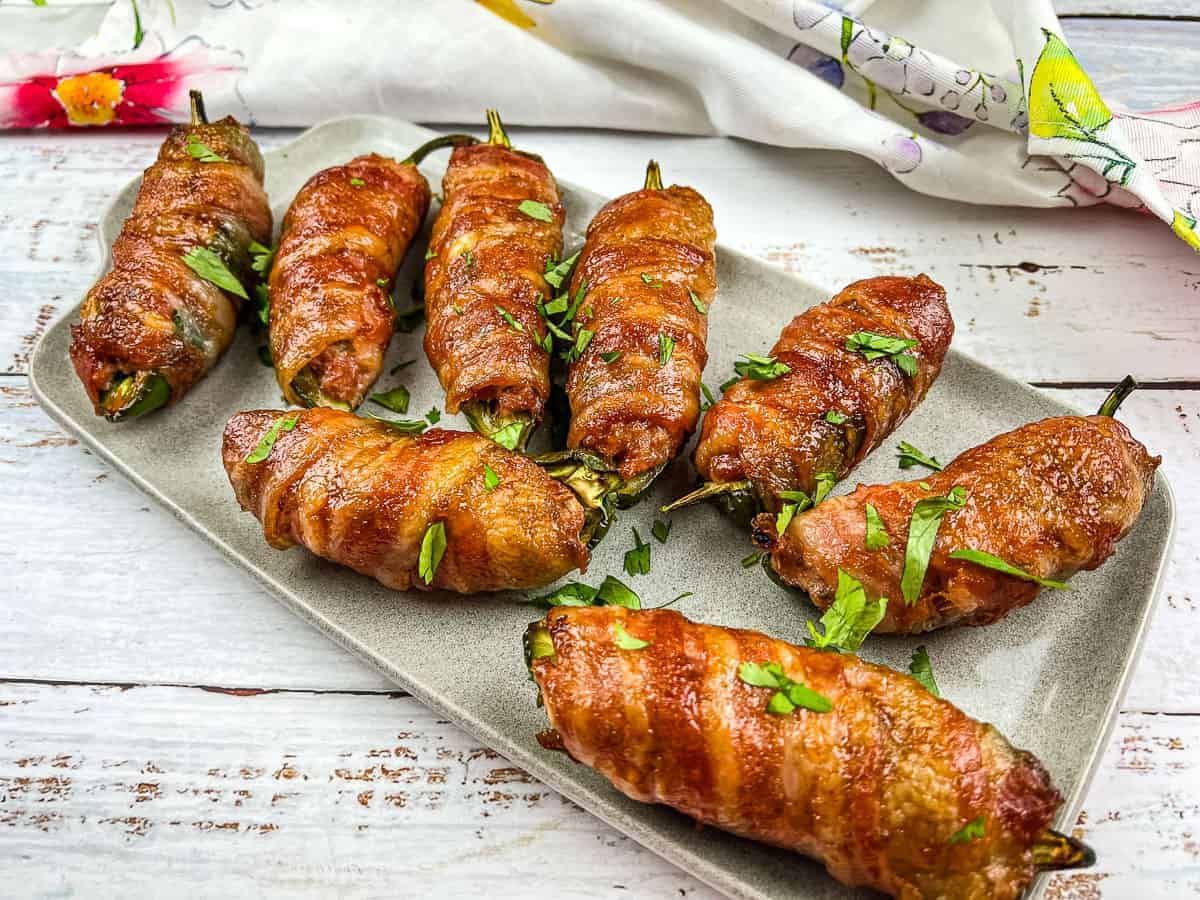 A rectangular plate holds eight bacon-wrapped jalape&ntilde;o poppers, garnished with chopped parsley, on a white wooden surface with a colorful napkin in the background.