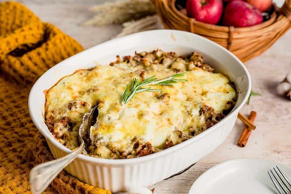A baked casserole topped with melted cheese and a sprig of rosemary sits in a white dish with a serving spoon, near a basket of red apples and a yellow cloth.