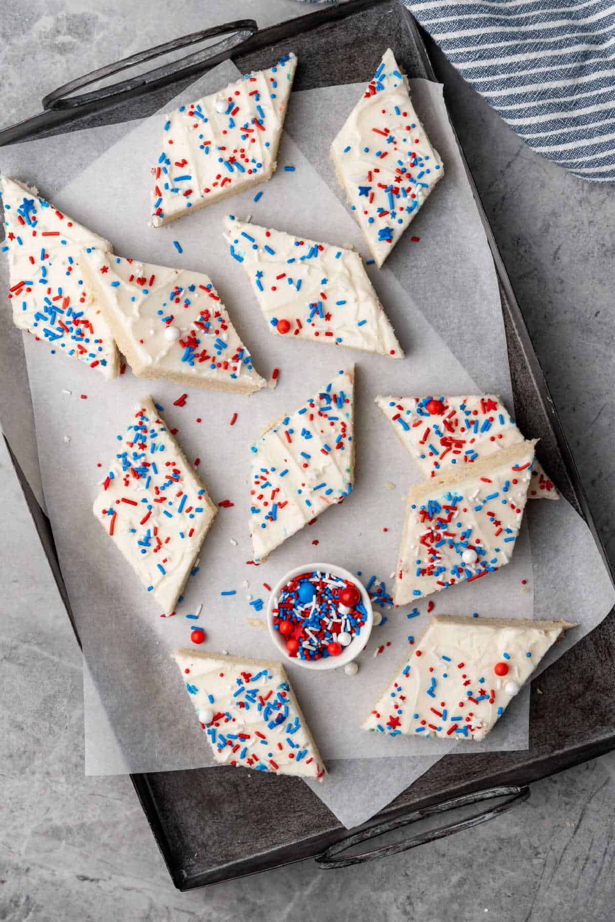 Iced sugar cookie bars cut into diamond shapes, topped with white frosting and red, white, and blue sprinkles, arranged on a parchment-lined tray with a small bowl of sprinkles.