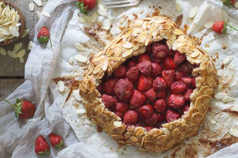 A rustic strawberry galette with sliced almonds on the crust sits on parchment paper, surrounded by fresh strawberries and almond slices.