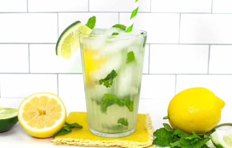 A glass of iced lemonade with mint leaves, a lime slice, and a striped straw is on a yellow coaster, surrounded by fresh lemon, lime, and mint against a white tiled background.