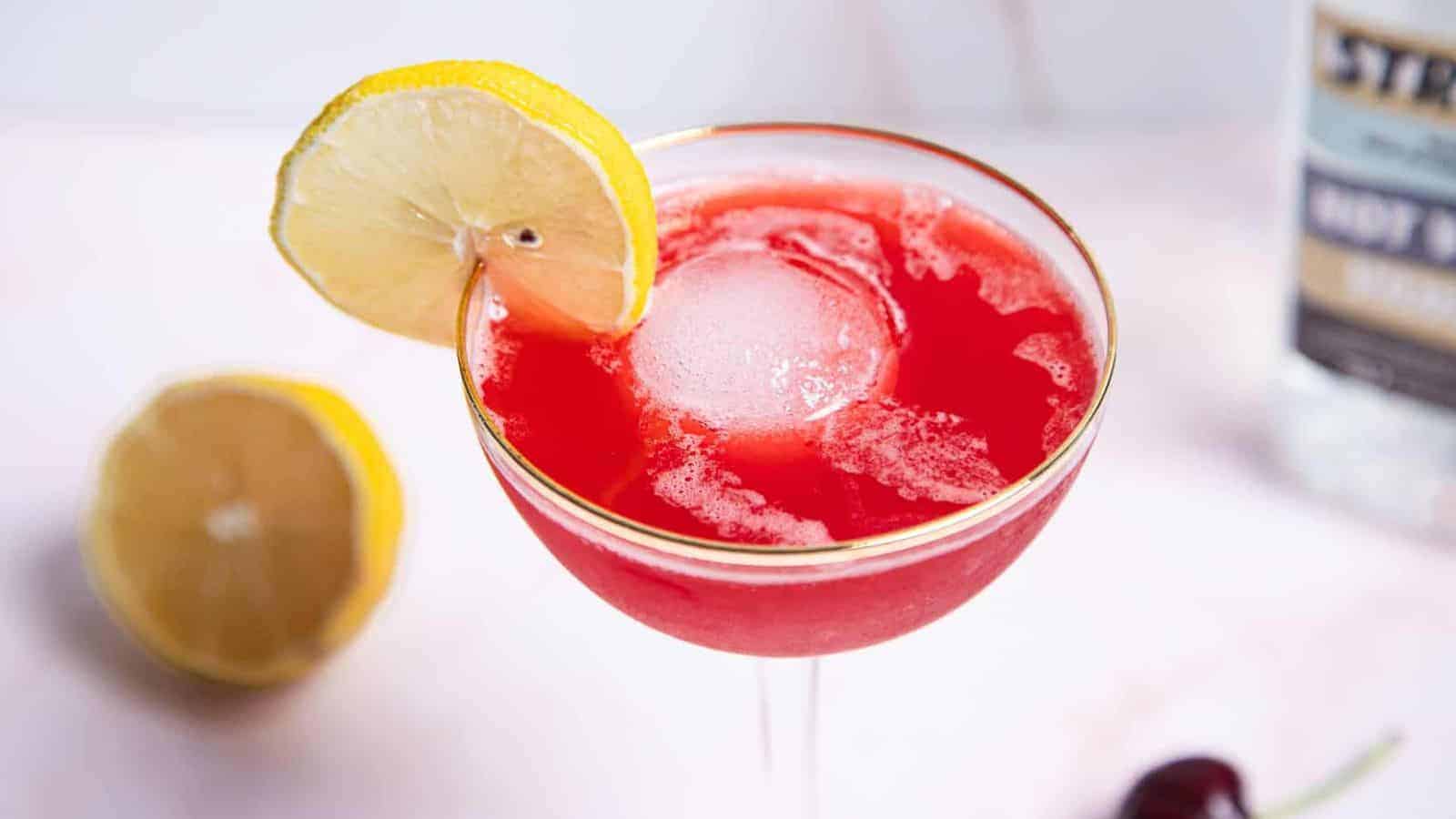 A red cocktail with a round ice cube is served in a coupe glass, garnished with a lemon slice. A halved lemon and a cherry are visible in the background.
