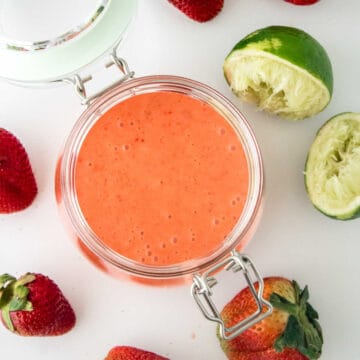A glass jar filled with strawberry sauce sits on a white surface surrounded by fresh strawberries and a squeezed lime.