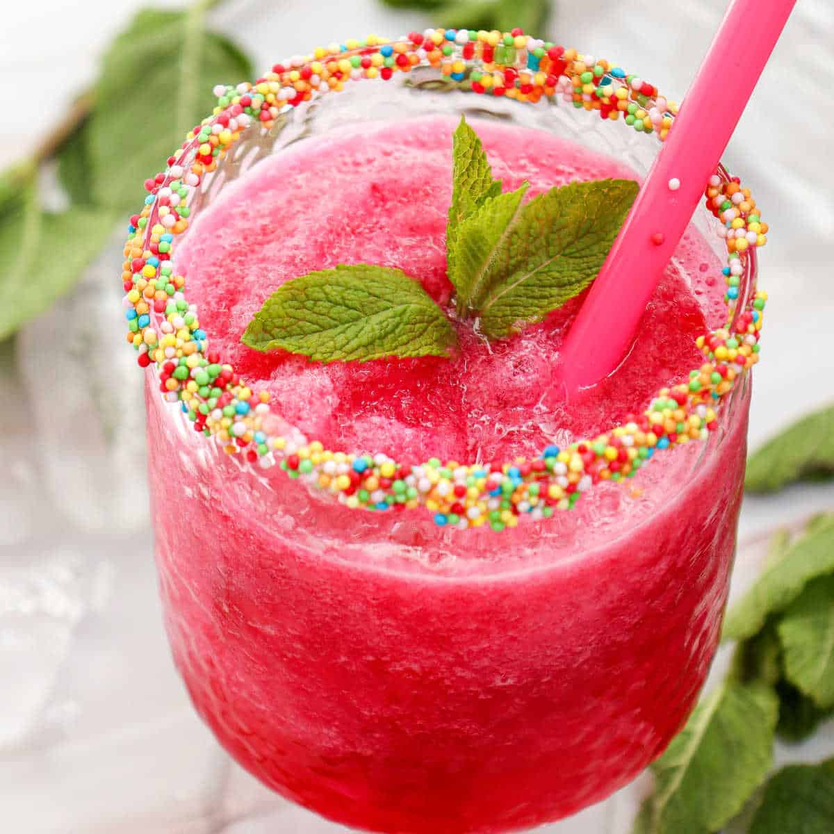 A pink slushy drink in a glass with a colorful sprinkle rim, garnished with mint leaves and served with a pink straw.
