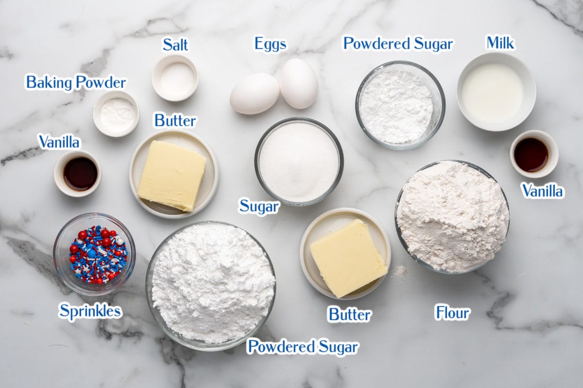 Various labeled baking ingredients including flour, sugar, butter, eggs, powdered sugar, milk, vanilla, baking powder, salt, and sprinkles arranged on a marble surface.