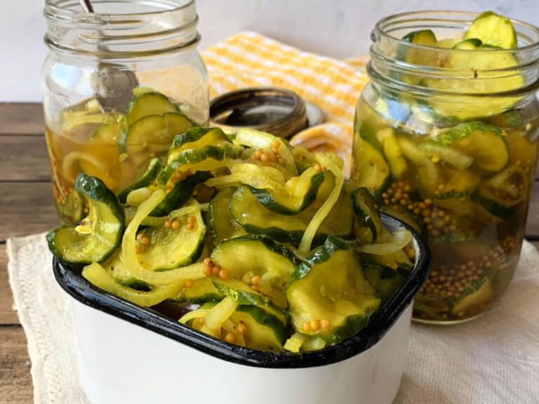 A white bowl and two glass jars filled with sliced pickles, onions, and mustard seeds sit on a cloth-covered wooden surface.