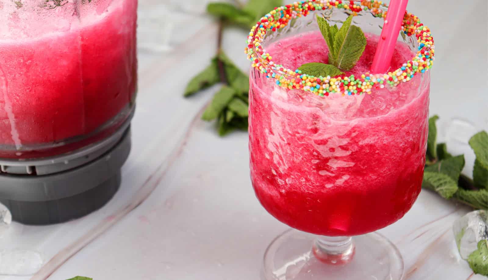A bright pink slush drink in a glass with a sprinkle-coated rim, garnished with mint leaves and a straw, sits next to a blender and fresh mint on a white surface.