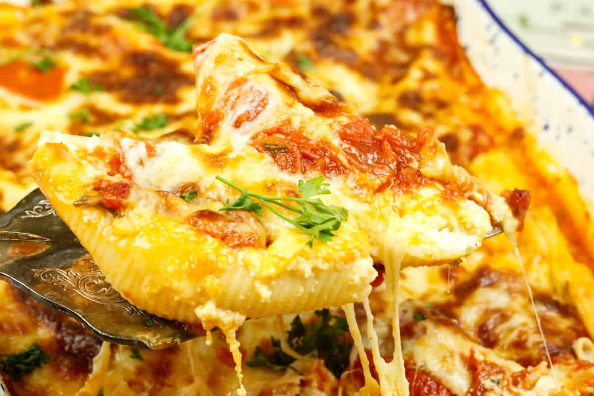 A close-up of a cheesy stuffed pasta shell topped with tomato sauce and parsley being lifted from a pan of baked pasta.
