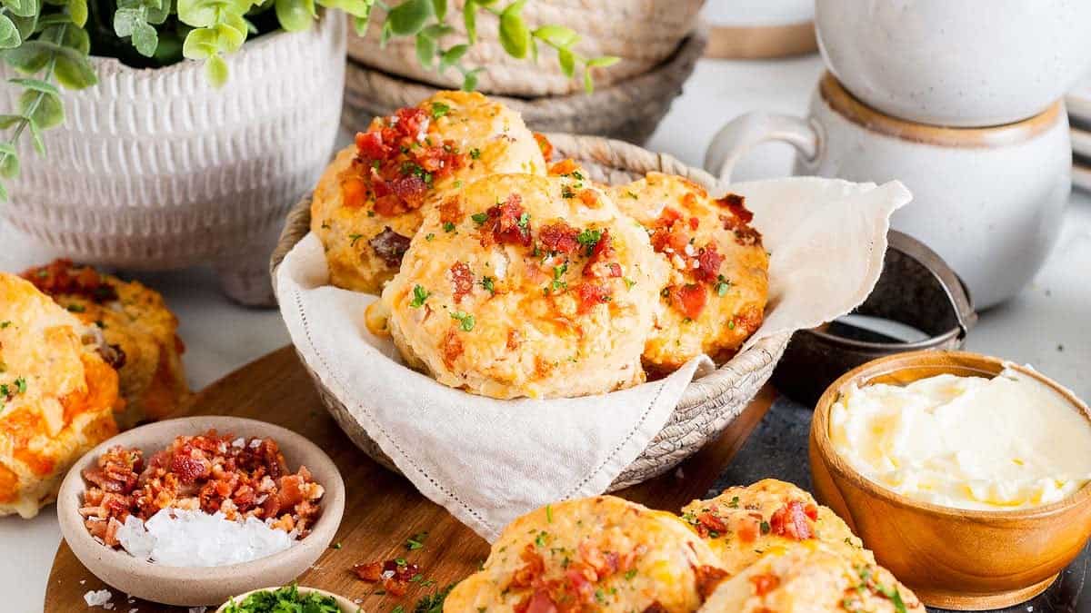 A basket of cheese and bacon biscuits garnished with herbs, surrounded by bowls of butter, chopped bacon, salt, and chopped herbs on a wooden surface.