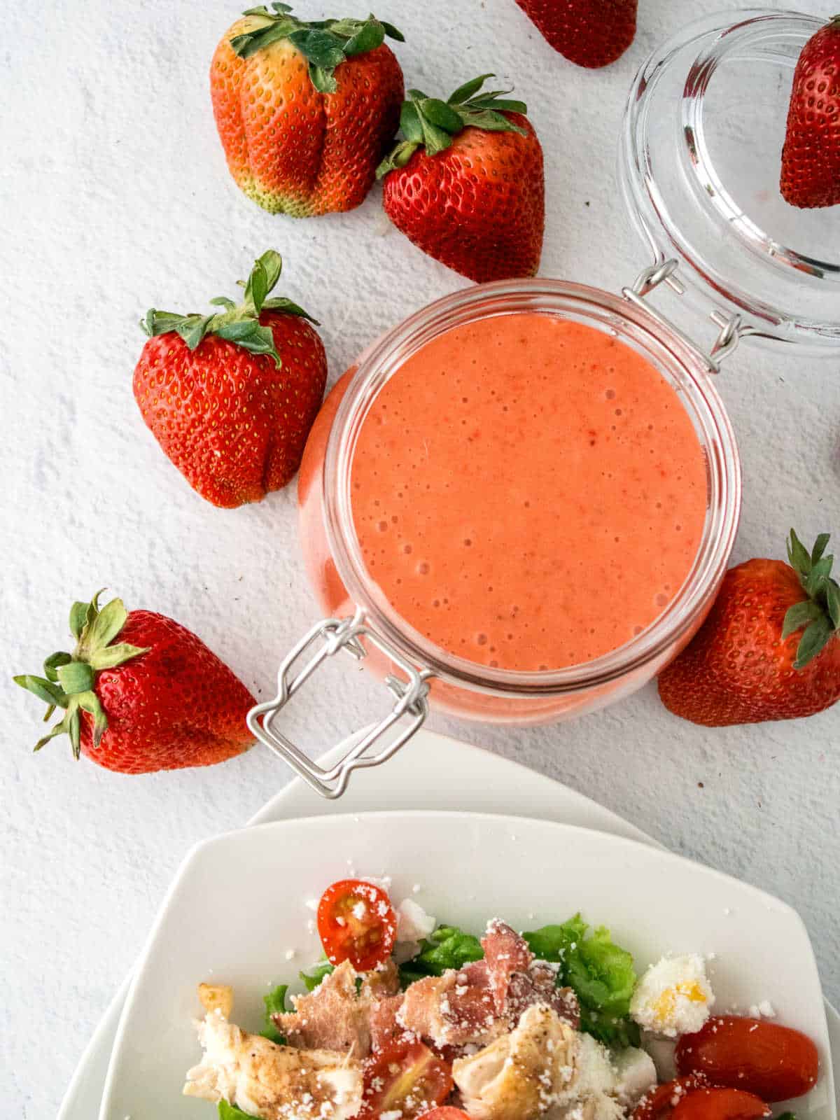 A jar of strawberry salad dressing surrounded by fresh strawberries sits next to a plate of salad with cherry tomatoes and cheese on a light surface.