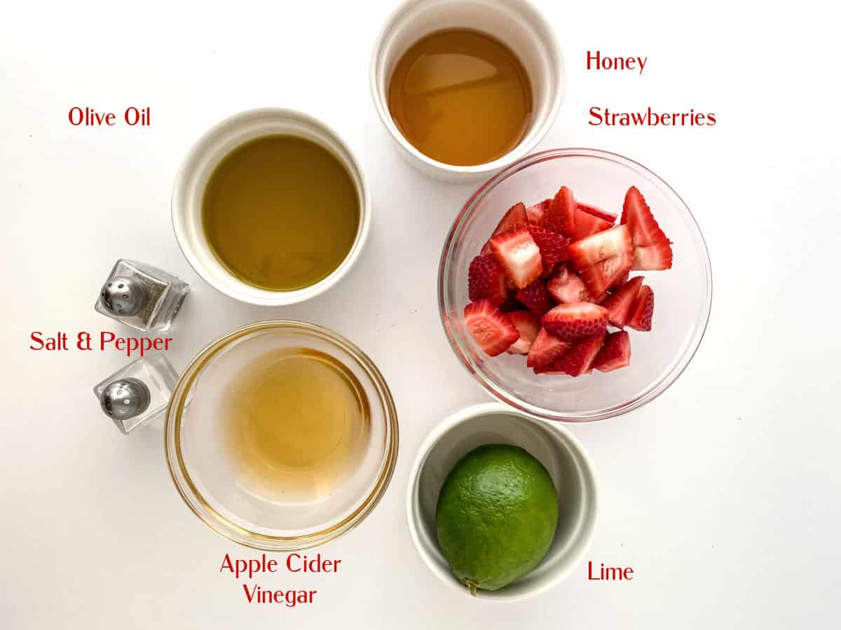 Top-down view of bowls containing olive oil, honey, apple cider vinegar, sliced strawberries, a whole lime, and salt and pepper shakers, each labeled with red text.