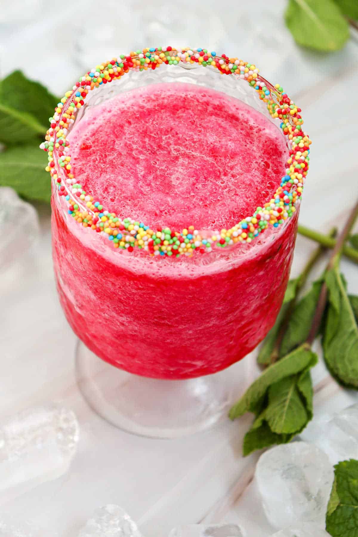 A bright pink slushy drink in a glass with a rim coated in colorful sprinkles, surrounded by ice cubes and fresh mint leaves.